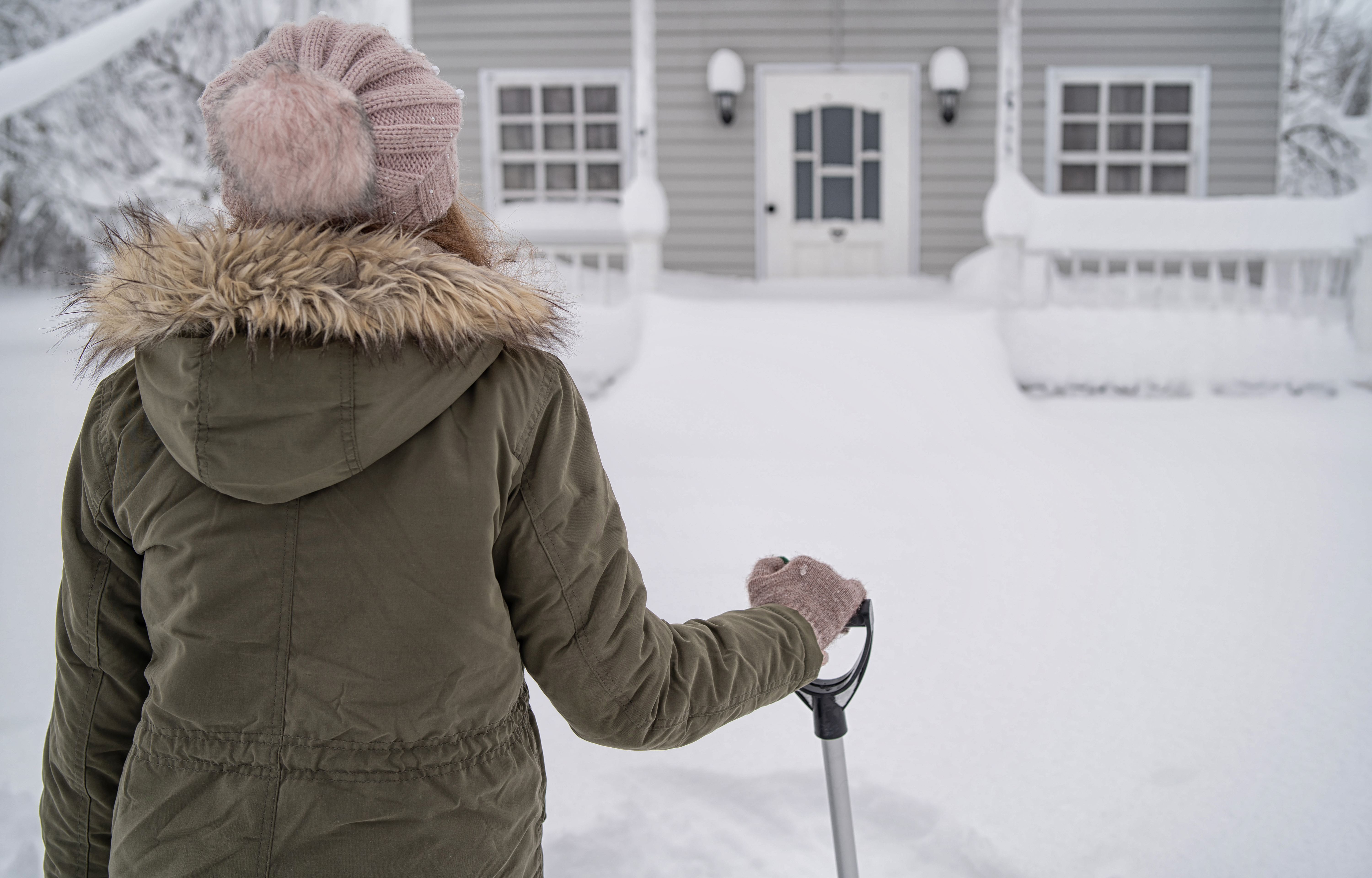 Woman in a winter hat and coat looking out over snowy scene in front of house.