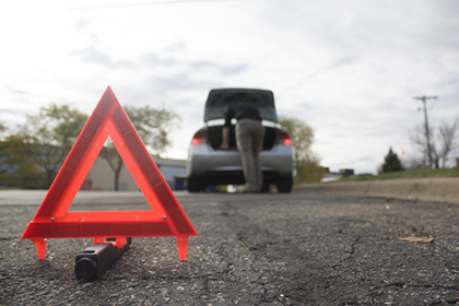 Red and orange triangle sign on road in foreground with person looking in car trunk in background.