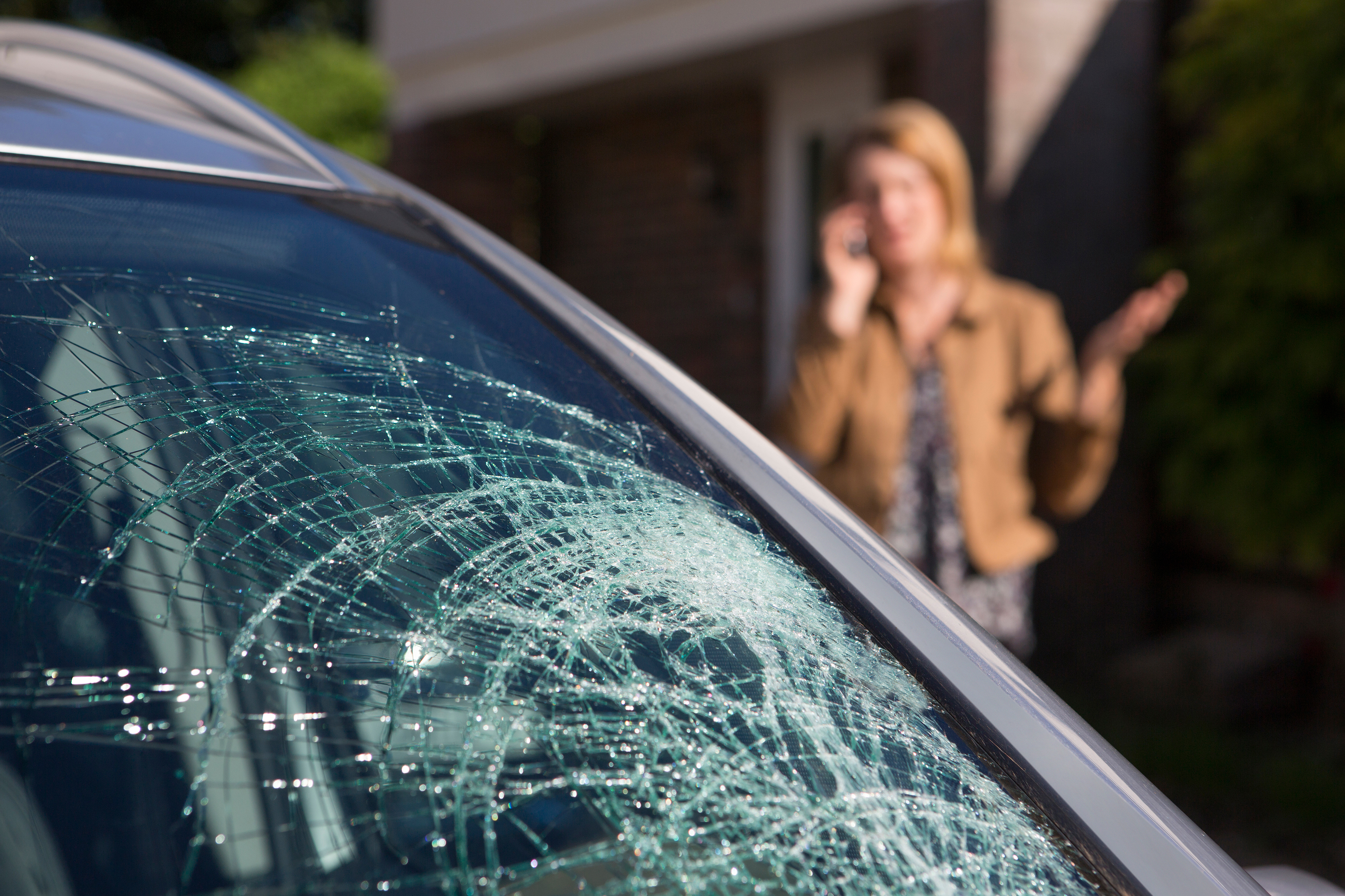 Close-up of vehicle with smashed windshield with woman on the phone in the background.